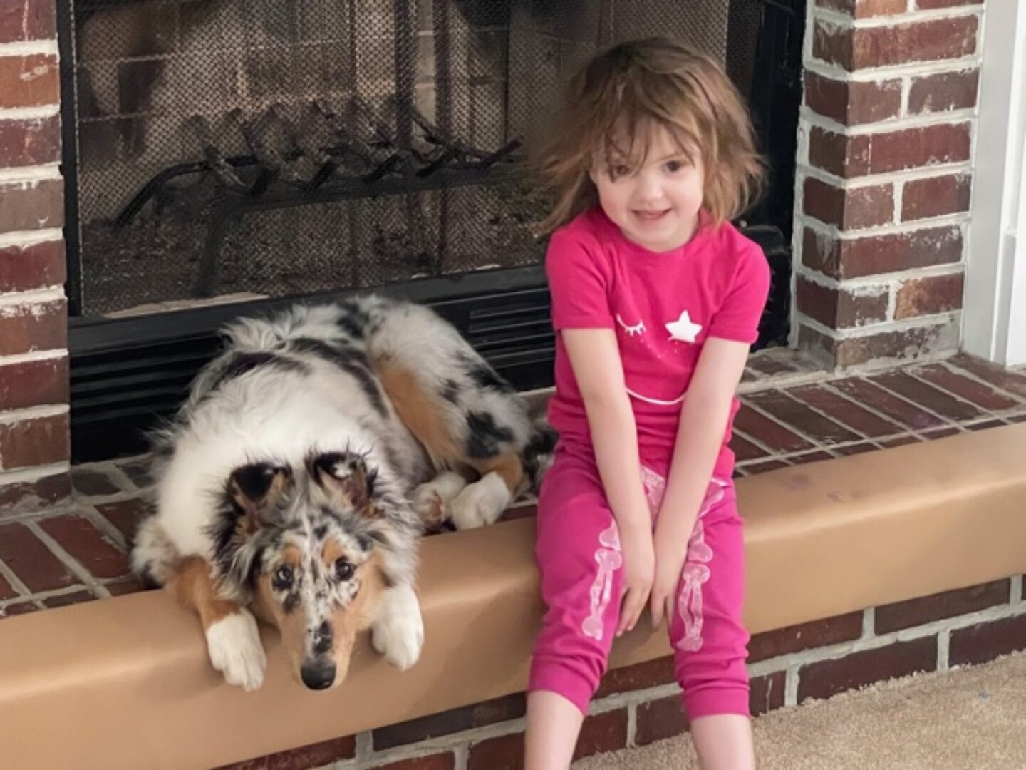 A girl and her dog sitting on the edge of a fireplace.